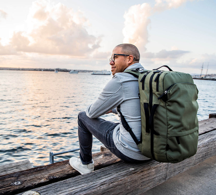 Man wearing glasses and a light gray shirt sitting on a wooden dock by the water at sunset, carrying a large green Travel Backpack