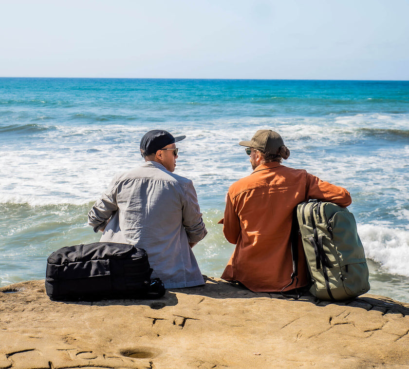 Two men sitting on a rock by the ocean, each with a Travel backpack beside them, facing the water under a clear sky.