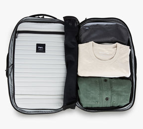Open Travel Backpack packed with folded clothes including a beige Everlane t-shirt and a green button-up shirt, and a black packing cube labeled 'Pakt.' inside a suitcase with a 'Pakt.' logo on the interior flap.