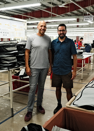 Two men standing side by side in a factory or workshop with tables and fabric materials around them.