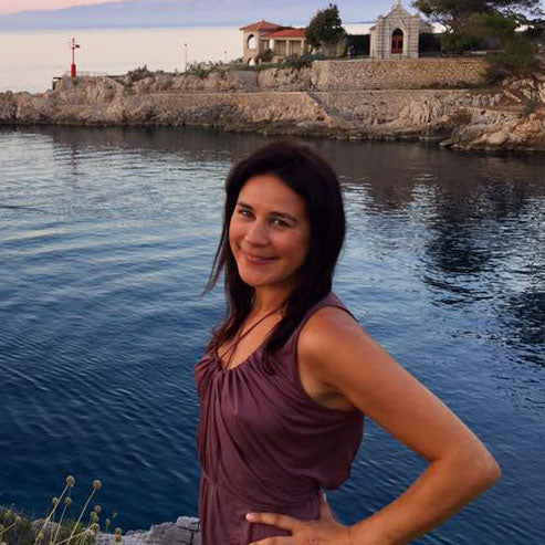 Woman in a purple sleeveless top standing by a rocky shoreline with calm water and buildings in the background.
