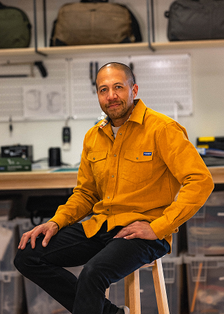 Pakt's founder wearing a mustard yellow shirt sitting on a stool in a design studio with tools and storage bins in the background.