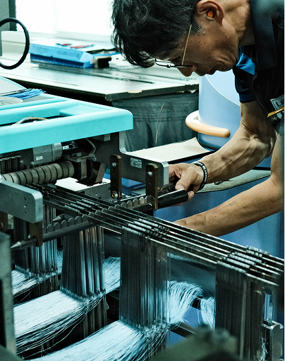 Man operating a textile weaving machine with threads running through the machine's metal parts in a workshop setting.