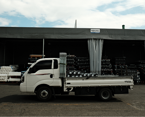 White flatbed truck parked in front of a warehouse with stacked rolls of material and metal pipes on the truck bed.