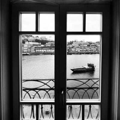 View through tall window and wrought-iron balcony railing to river with a small boat and buildings on the opposite bank