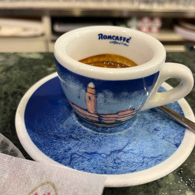 Espresso in a blue Romcaffè caffetteria cup and matching saucer with lighthouse design, spoon on saucer.