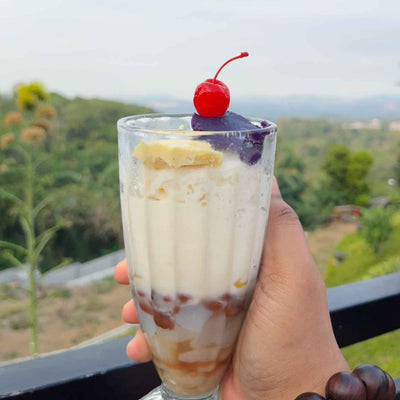 Hand holding layered dessert glass with red cherry, purple ube scoop, banana slice and beans, hillside view background.