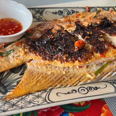 Grilled whole fish garnished with chili and herbs on a decorative plate, served with a small bowl of dipping sauce.