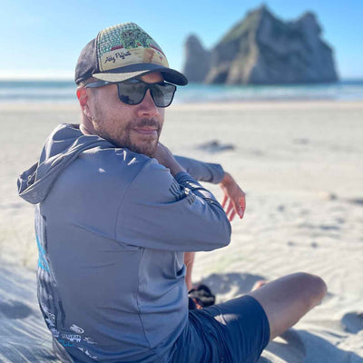 Man sitting on a sandy beach wearing sunglasses and a cap, with a rocky island visible offshore.