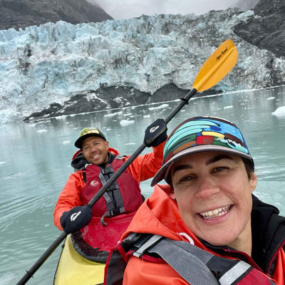 Two smiling kayakers in orange jackets taking a selfie in a yellow tandem kayak with glacier and ice chunks behind them.