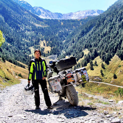 Motorcyclist in neon green jacket standing beside a loaded adventure motorcycle on a rocky mountain dirt road