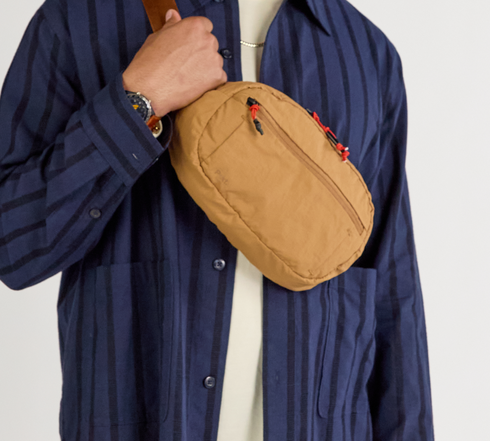 Mojave-colored Stash Sling worn crossbody over navy striped shirt; tan bag with red zipper pulls visible.