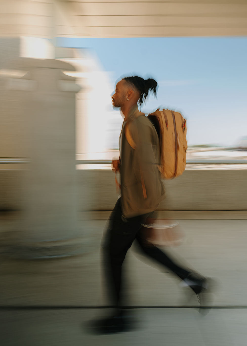 Blurred side view of a person walking quickly with a tan backpack under a covered walkway