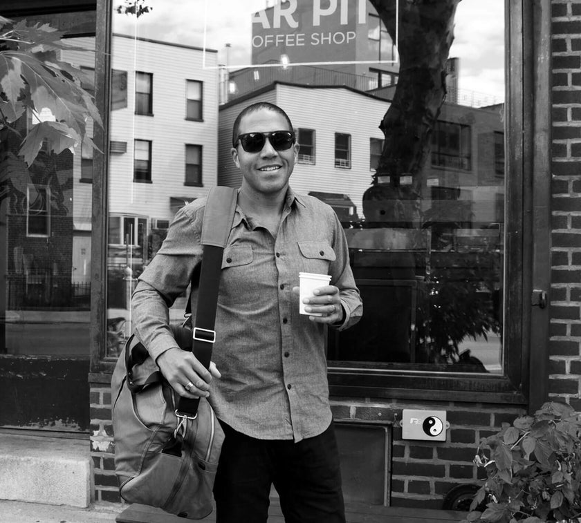 Man wearing sunglasses, holding a coffee cup and carrying a shoulder bag, standing outside a coffee shop with a sign reading 'AR PIT COFFEE SHOP' visible in the window.