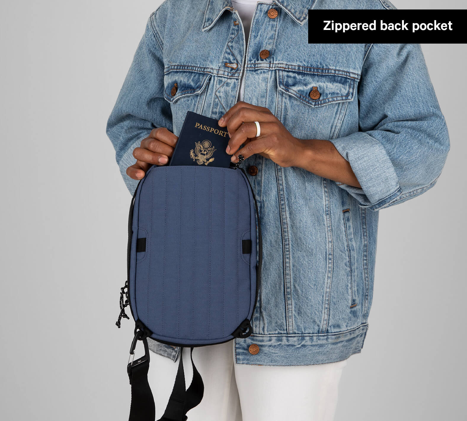 Person wearing a denim jacket holding a blue Everyday 5L Sling bag with a passport partially inside the zippered back pocket labeled 'Zippered back pocket'