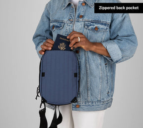 Person wearing a denim jacket holding a blue Everyday 5L Sling bag with a passport partially inside the zippered back pocket labeled 'Zippered back pocket'