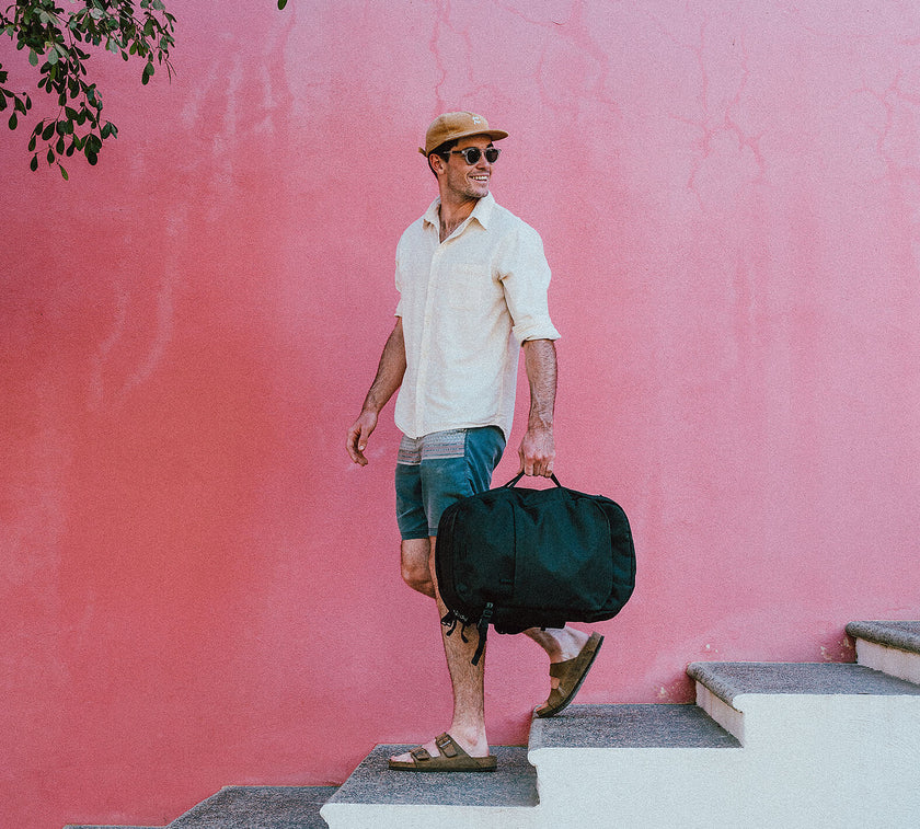 Man wearing sunglasses, a tan cap, light shirt, and shorts, carrying a black duffel bag while walking down stairs against a pink wall.