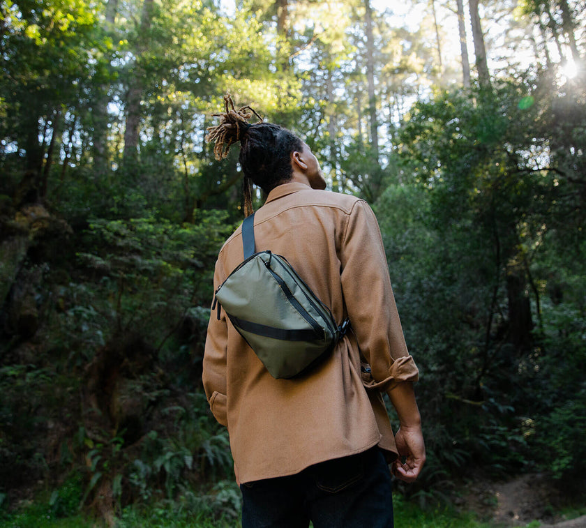 Person with dreadlocks wearing a tan jacket and green Anywhere 5L Sling looking up in a sunlit forest