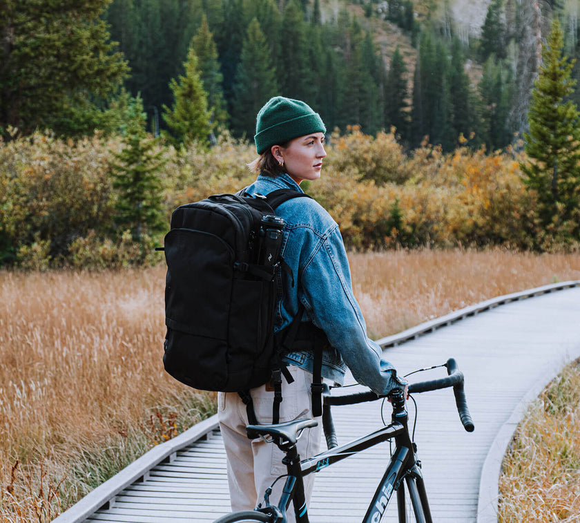 Person wearing a green beanie and denim jacket holding a black bicycle on a wooden path surrounded by autumn foliage and trees.