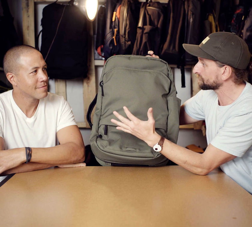 Two men sitting at a table, one holding and showing a green V1 Travel Backpack to the other in a store or workshop setting with backpacks hanging in the background.