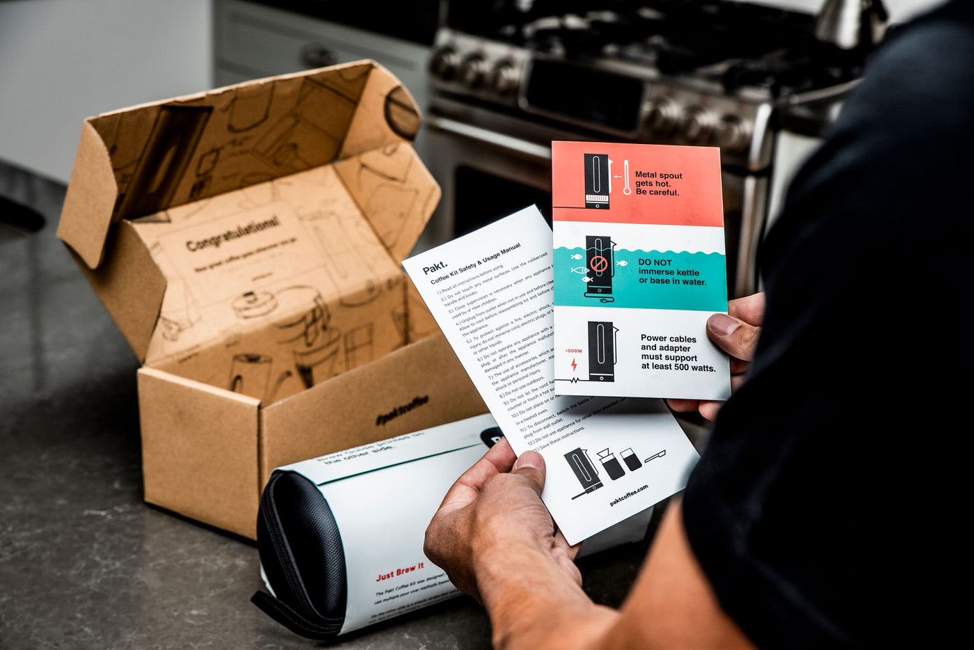 Person holding instruction manuals for a coffee kit, showing safety warnings: "Metal spout gets hot. Be careful.", "DO NOT immerse kettle or base in water.", "Power cables and adapter must support at least 500 watts.", and the website "paktcoffee.com".