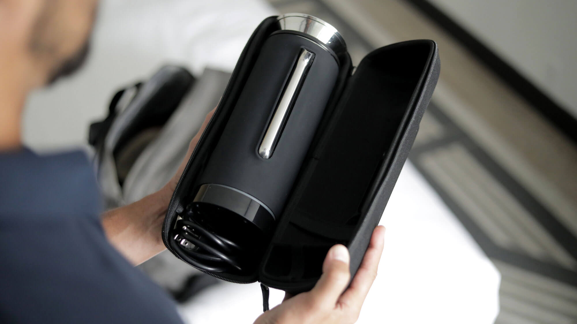 Person holding the V1 Grandcart Coffee Kit inside a black zippered carrying case, with a visible power cord coiled inside the case.
