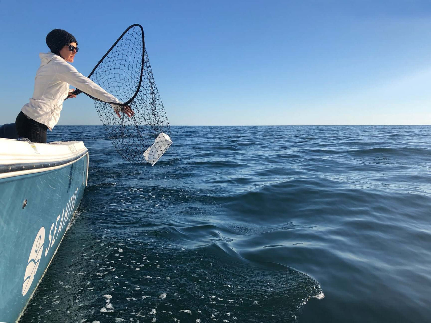 Person on a boat using a net to remove a floating plastic container from the ocean surface during a clear day.