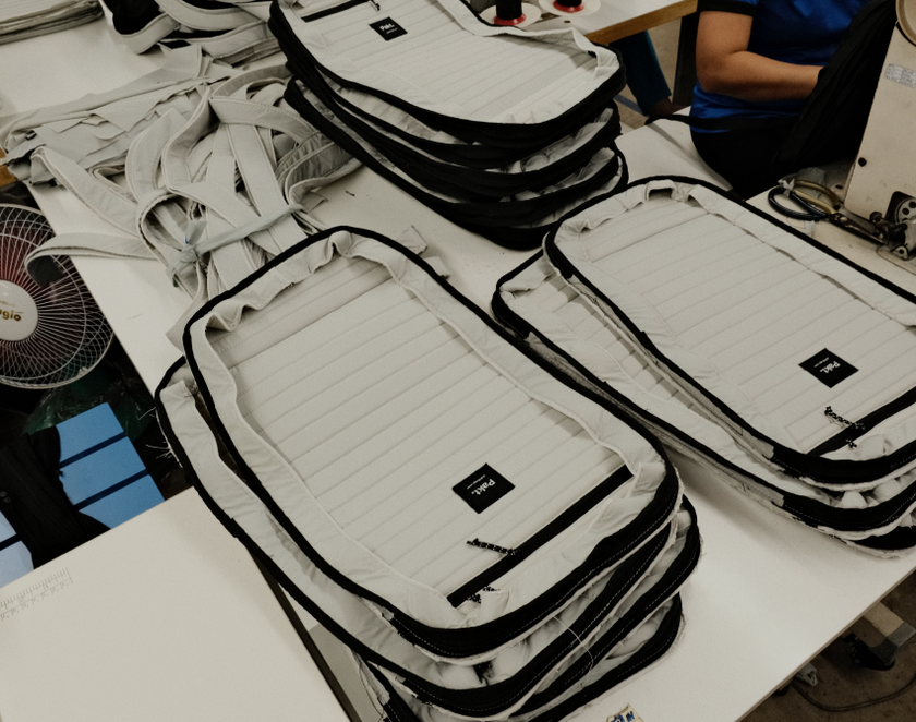 Stacked white padded bags with black trim labeled 'Peak Design' on a table in a workshop setting with a person sewing in the background.