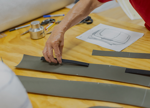 Person arranging strips of material on a wooden table with a sketch of a car design and tools in the background.
