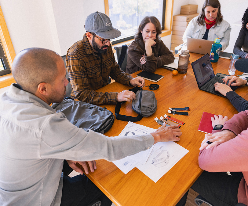 A group of six people sitting around a wooden table reviewing product samples and sketches, including watch straps and a small bag, in a meeting room with large windows.