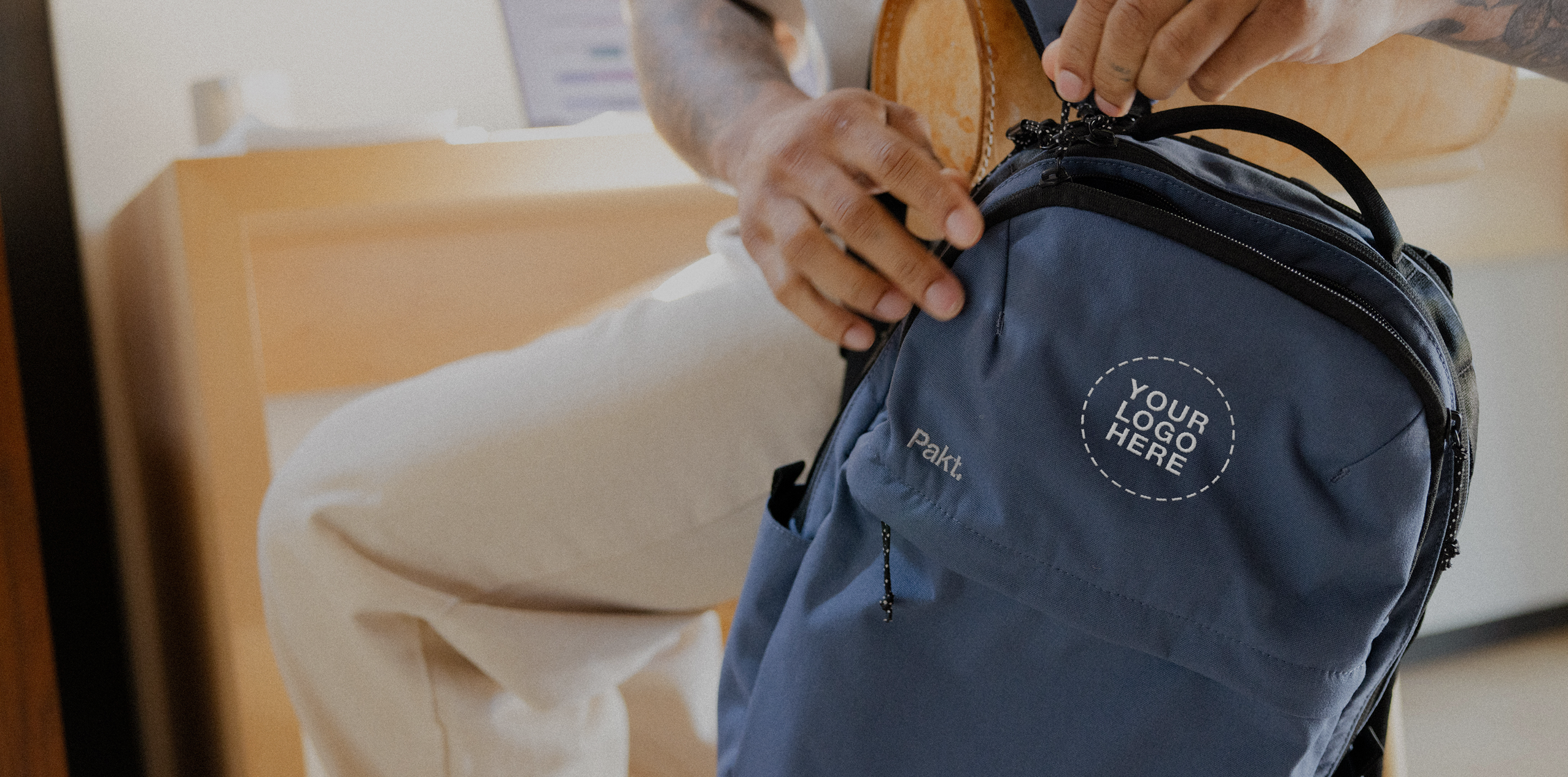 Navy backpack with circular patch reading "YOUR LOGO HERE" and small Grandcart. label; hands adjusting the top zipper.