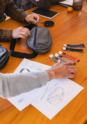 Hands adjusting a gray crossbody bag strap on a table with various watch straps and design sketches scattered around.