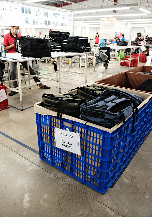 Backpacks in a blue crate labeled 'HANG BAY PASSED QC0006' inside a factory or warehouse with workers and tables in the background.