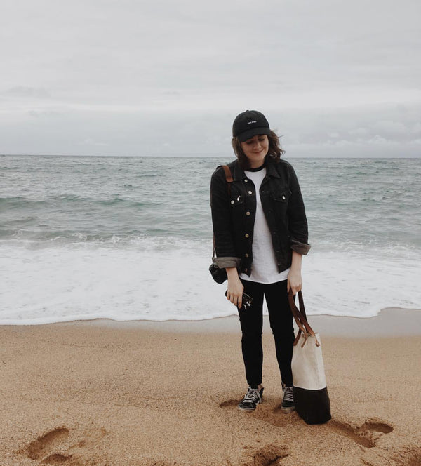 A person wearing a black jacket, black cap, and black pants standing on a sandy beach near the ocean with waves in the background, holding a two-tone tote bag.