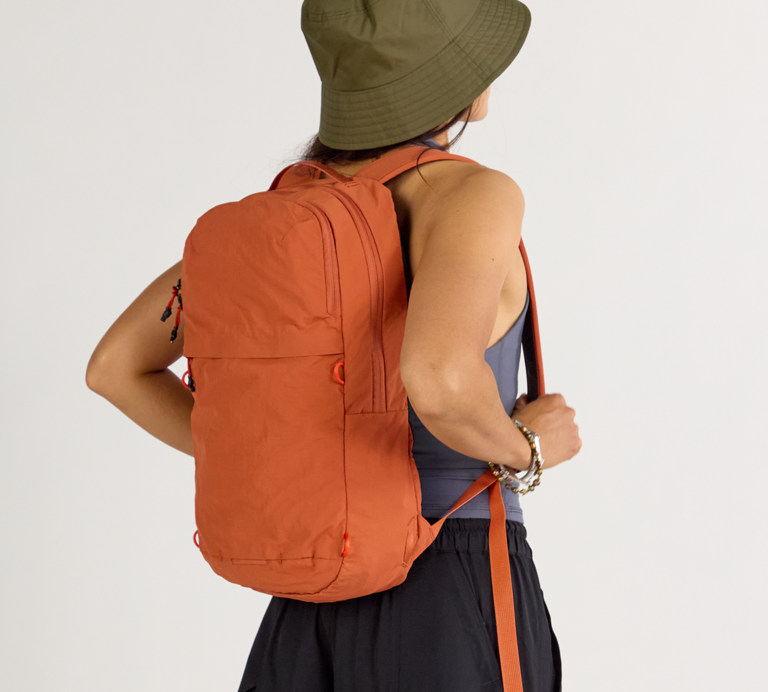 Back view of person wearing a rust-orange backpack and olive-green bucket hat against a plain background
