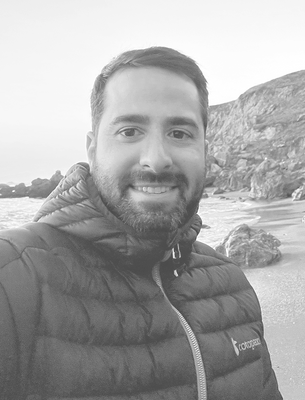 Smiling bearded person wearing a quilted jacket standing on a rocky beach with waves and cliffs behind.
