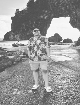 Person wearing floral shirt and sunglasses standing on rocky shore in front of a sea arch