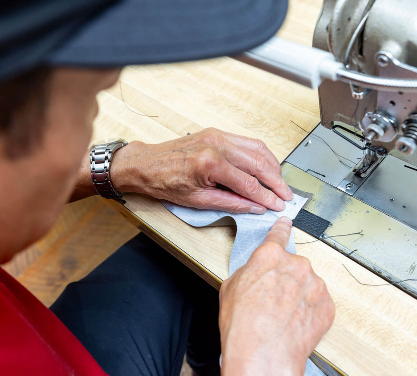 Person sewing fabric by hand near a sewing machine on a wooden table, wearing a watch and a blue cap.