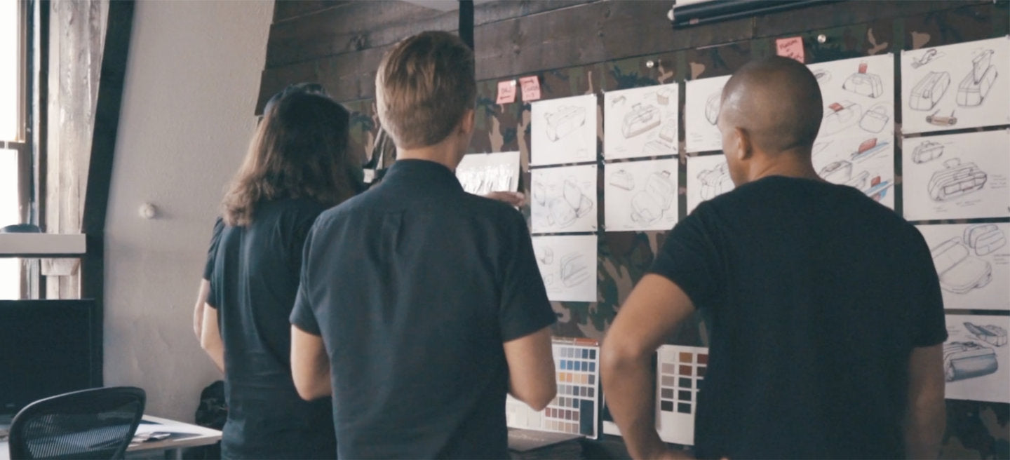 Three people standing in front of a wall covered with sketches and color swatches, discussing design ideas.