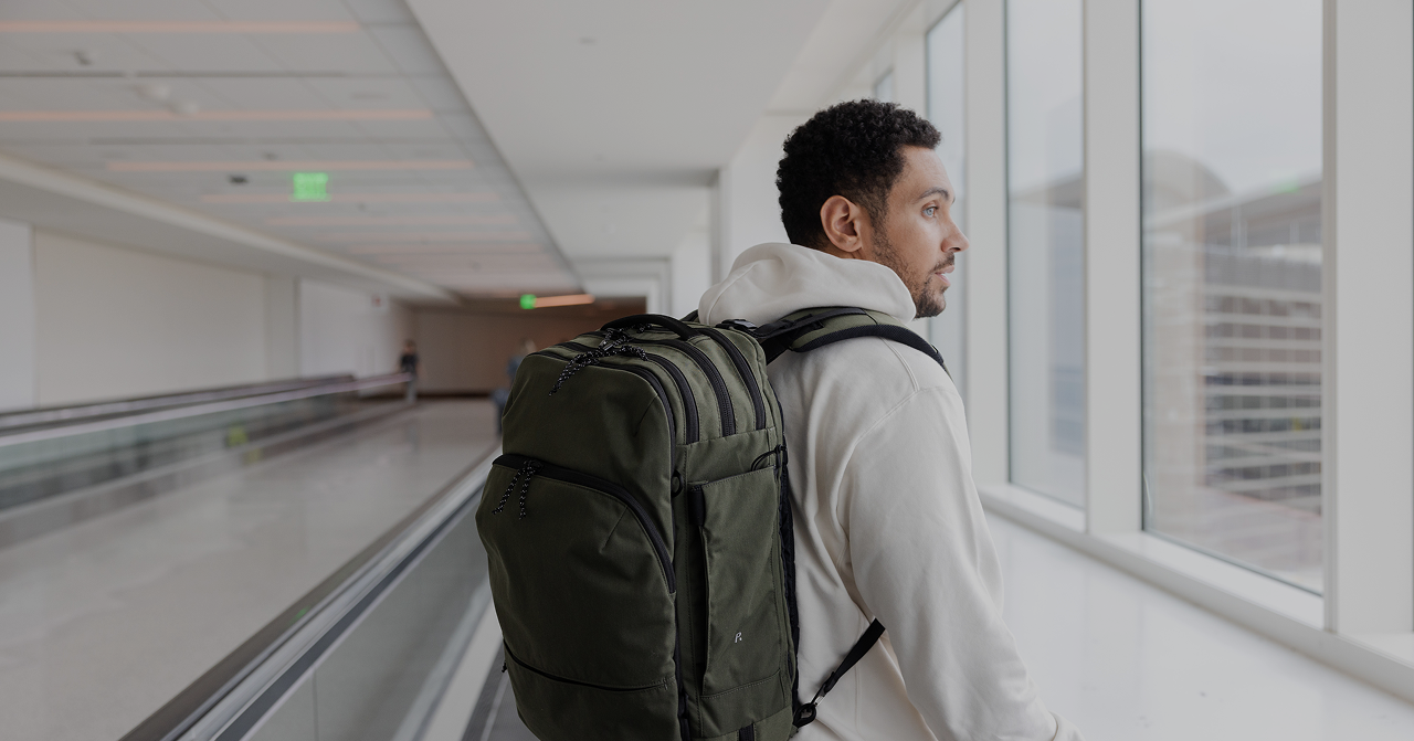 Man wearing a white hoodie and large green backpack looking out a window in an airport terminal