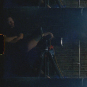 Person adjusting a camera on a tripod in a dimly lit room with a brick wall background. A behind the scenes photo.