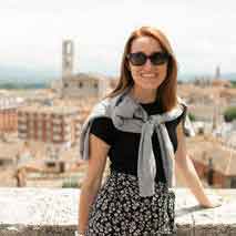 Smiling woman wearing sunglasses and a sweater draped over her shoulders on a stone ledge with a cityscape behind her