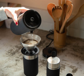 Pouring hot water from a black kettle into a coffee dripper over a black Grandcart Coffee Kit container on a kitchen counter with wooden utensils in the background