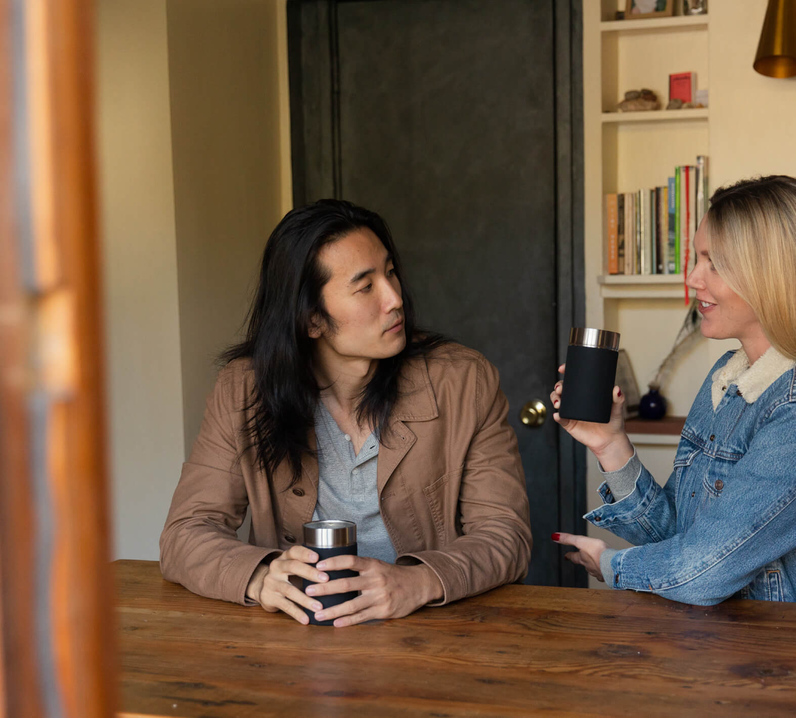 Two people sitting at a wooden table, each holding a coffee cup, engaging in conversation in a cozy room with bookshelves in the background.