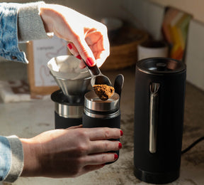 Person scooping ground coffee into a black cylindrical coffee grinder on a kitchen counter with a coffee dripper in the background
