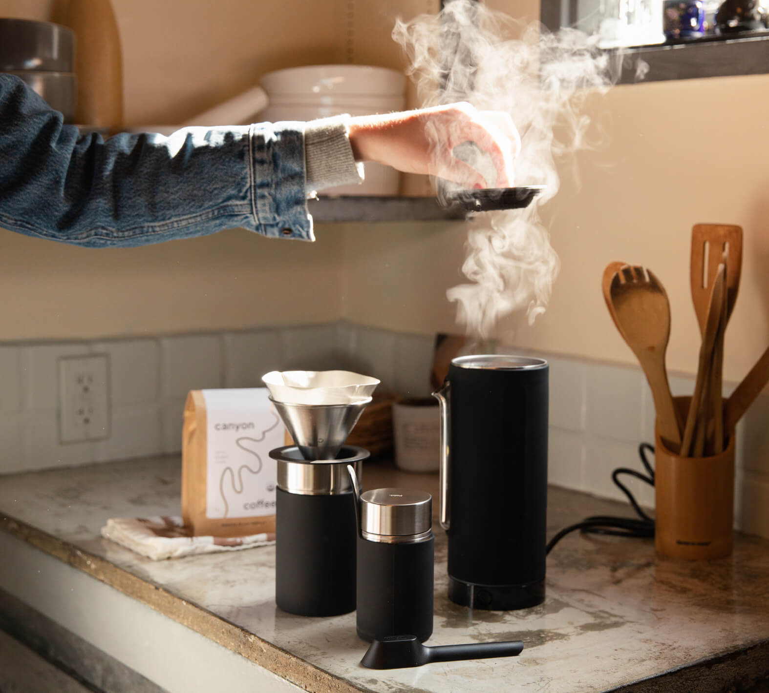 A hand holding a steaming cup above a coffee brewing setup with a dripper, grinder, and kettle on a kitchen counter.