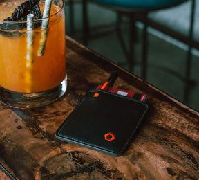Black Mojito Wallet with a red card partially visible, placed on a wooden table next to a glass with an orange drink and a straw.